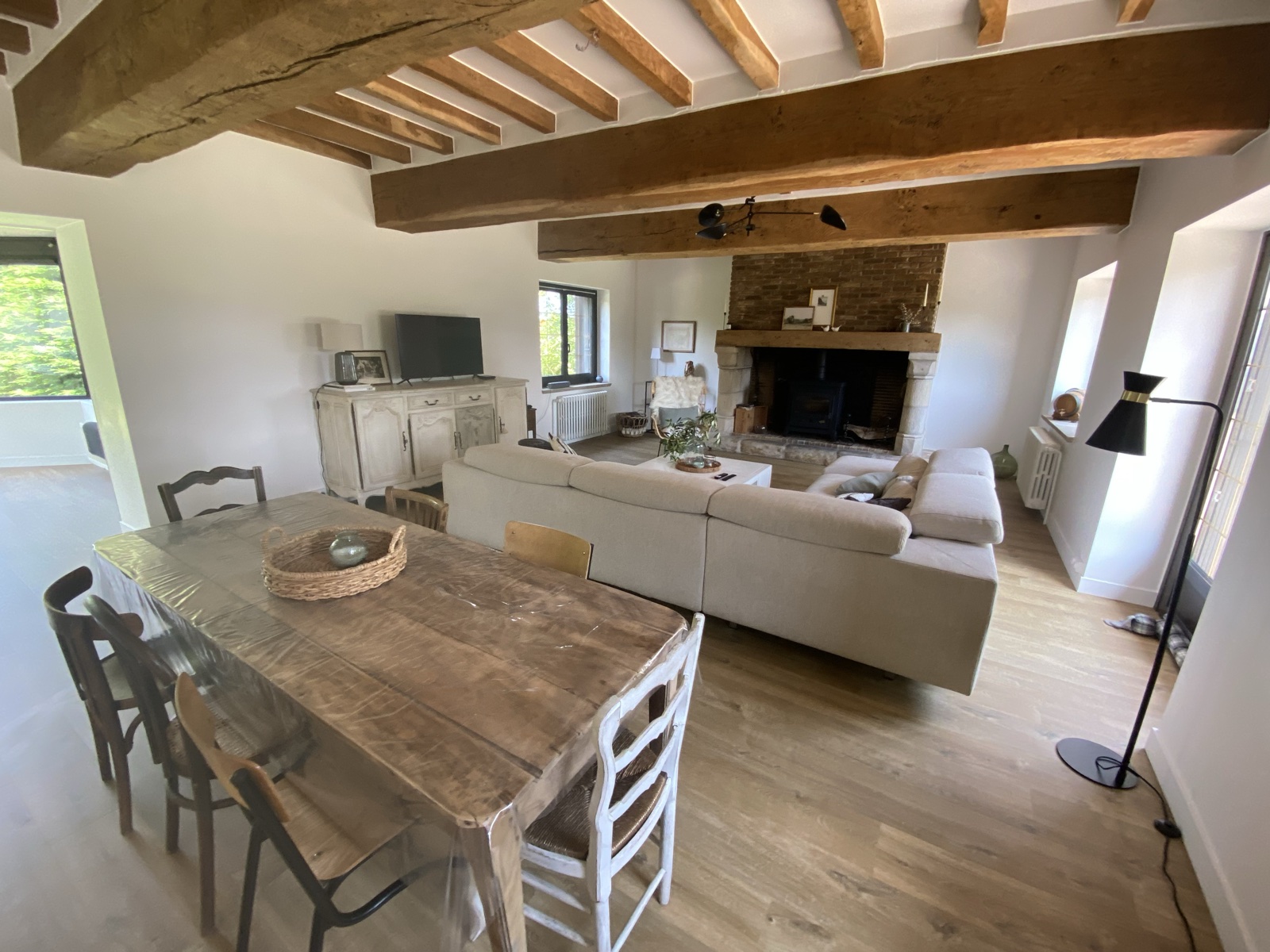 Open-plan salon with stone fireplace, wood stove, sofa and dining table beneath original Norman beams