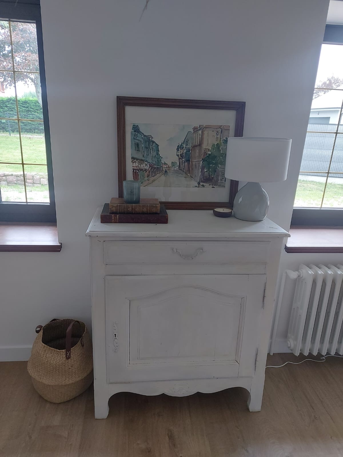 A white-painted Norman commode with watercolour, vintage books and table lamp between two windows
